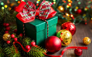 Green holiday gift box with red plaid bow, surrounded by Christmas ornaments and festive lights