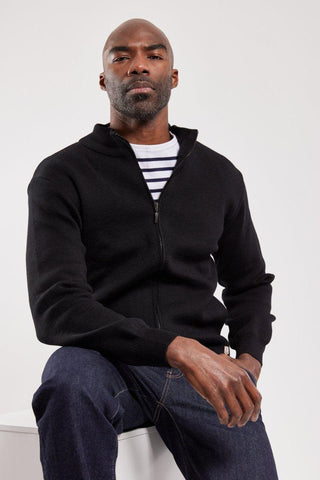 Man wearing black zip-up wool cardigan over striped shirt and dark jeans, seated indoors