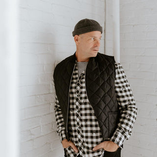 Man wearing grey wool beanie, black quilted vest, and plaid shirt against white brick wall