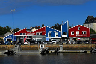 Colorful waterfront buildings with marine clothing stores, parked cars, and small boats in front.
