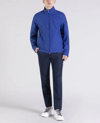 Man wearing blue nylon zip-up jacket, dark trousers, and white sneakers on white background