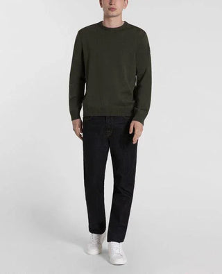 Man wearing olive green crew neck sweater, dark jeans, and white sneakers standing against white background