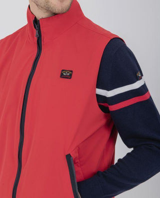 Red nylon sleeveless vest with zip and logo patch, worn over navy sweater with stripes