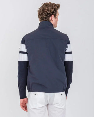 Man wearing navy sleeveless nylon vest, white pants, and navy sweater with white stripes, rear view
