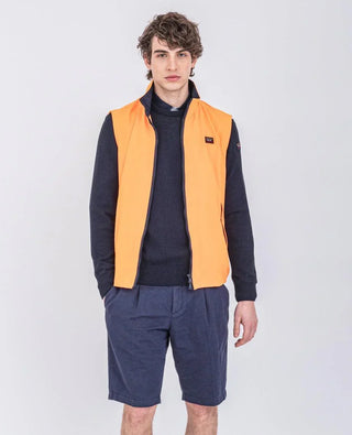 Man wearing orange nylon sleeveless vest, navy sweater, and navy shorts, standing indoors