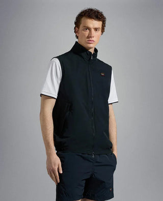 Man wearing navy nylon sleeveless vest over white shirt and navy shorts, standing indoors